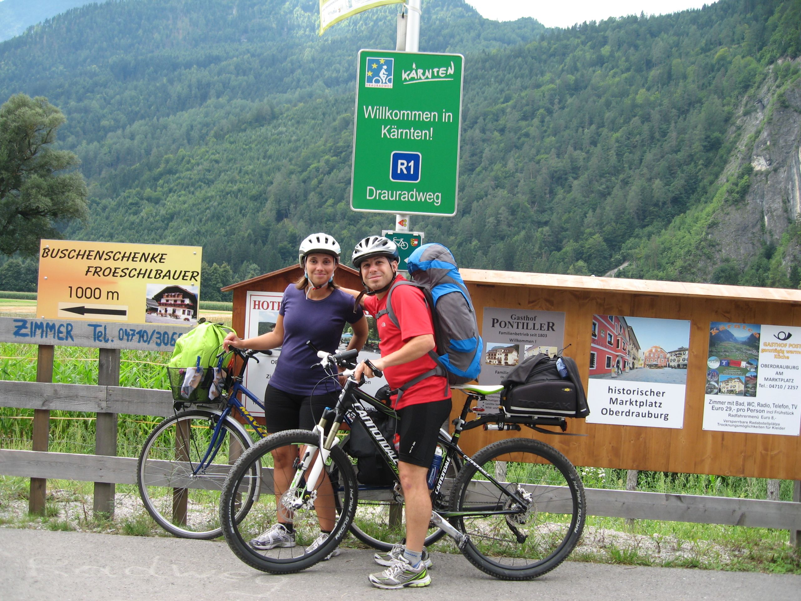 Due ciclisti in vacanza con "Girolibero" si fermano vicino a un cartello di confine lungo il percorso della Ciclabile della Drava.